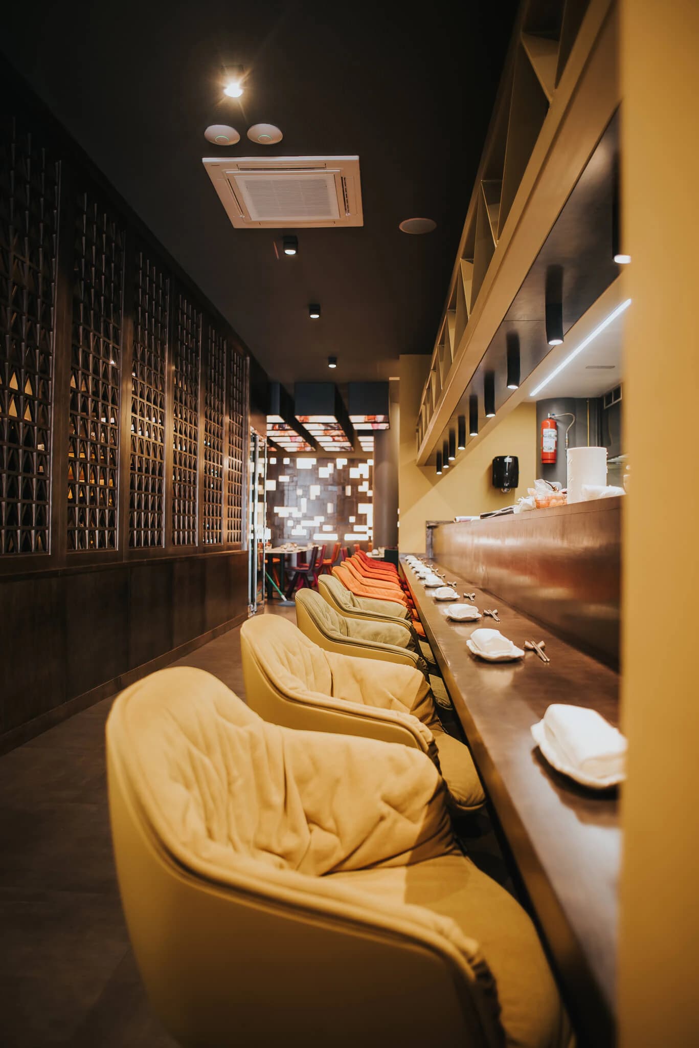 Mustard upholstered chairs along a narrow dining bar lit by warm shelving at Jaxana