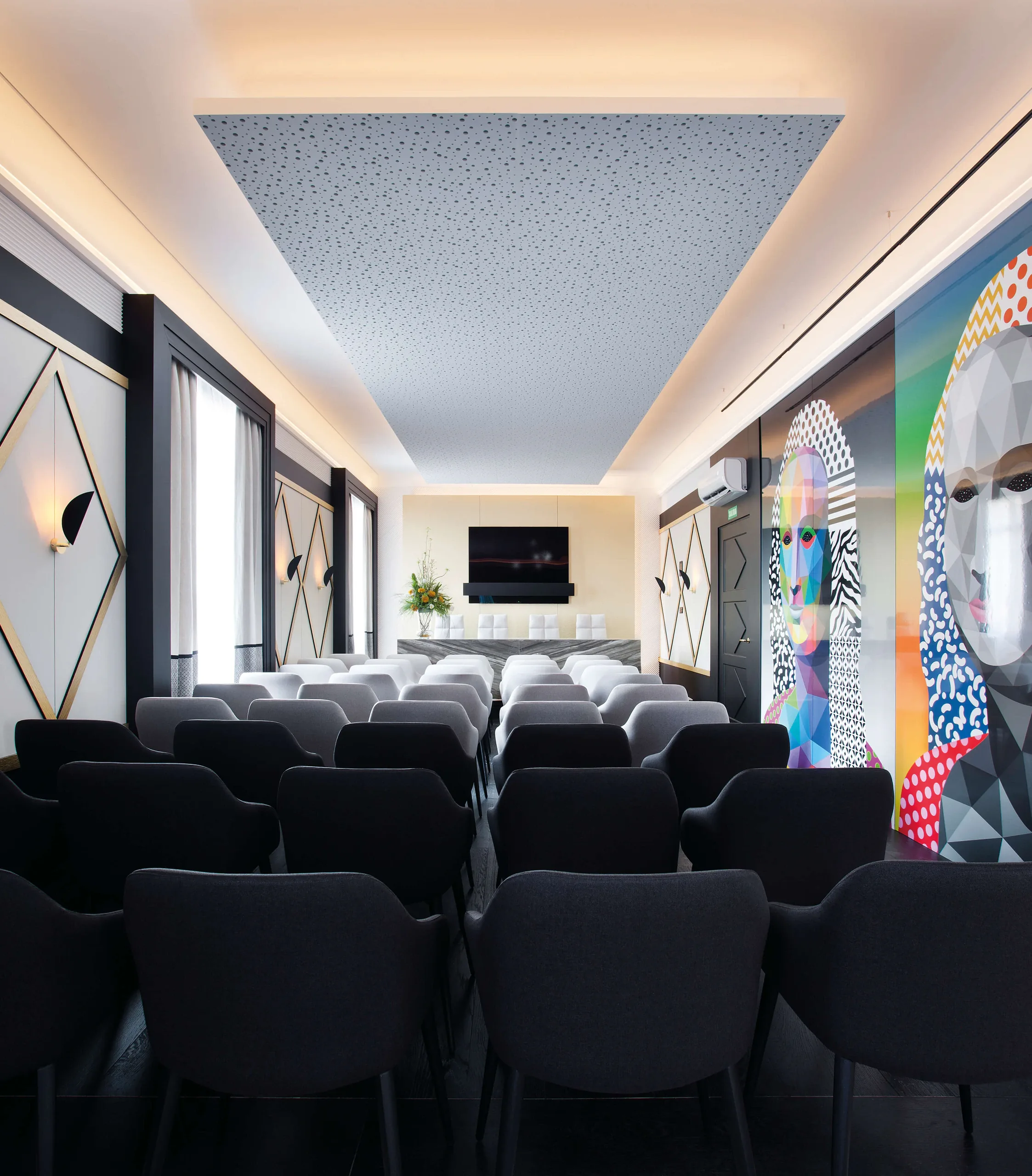 Rows of dark upholstered chairs in a softly lit auditorium with a patterned wall at Casa Decor 2019