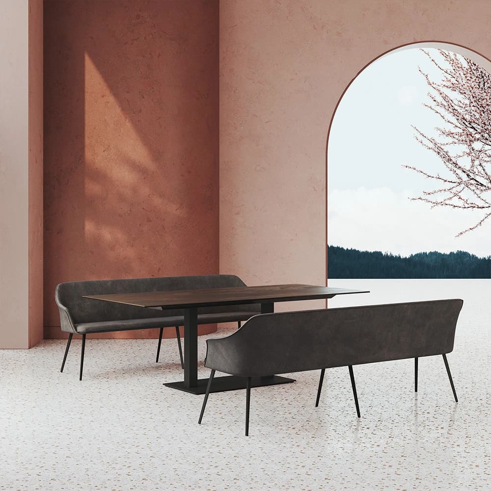 Dado dining table with a central metal base and upholstered benches beside a terracotta wall and arched window