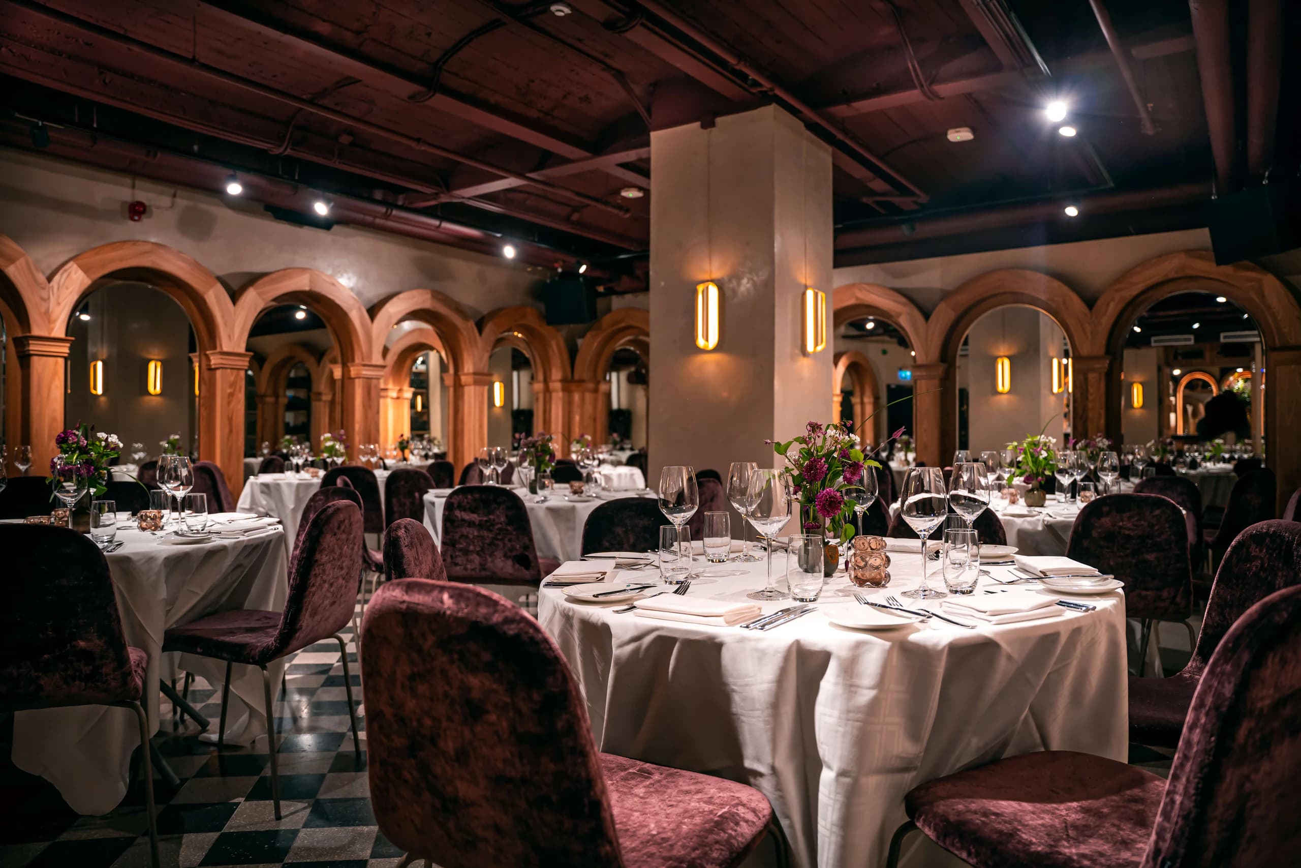 Plum upholstered chairs and white-clothed tables beneath warm timber arches at Bank Hotel