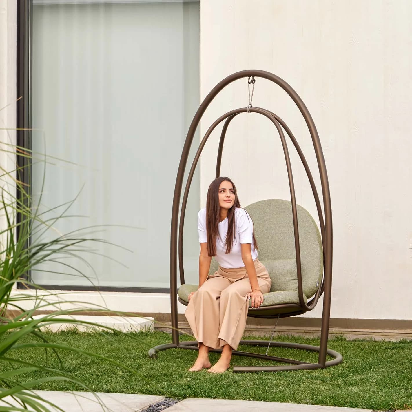 A woman sitting in a Niu swing chair on a lawn beside a white house
