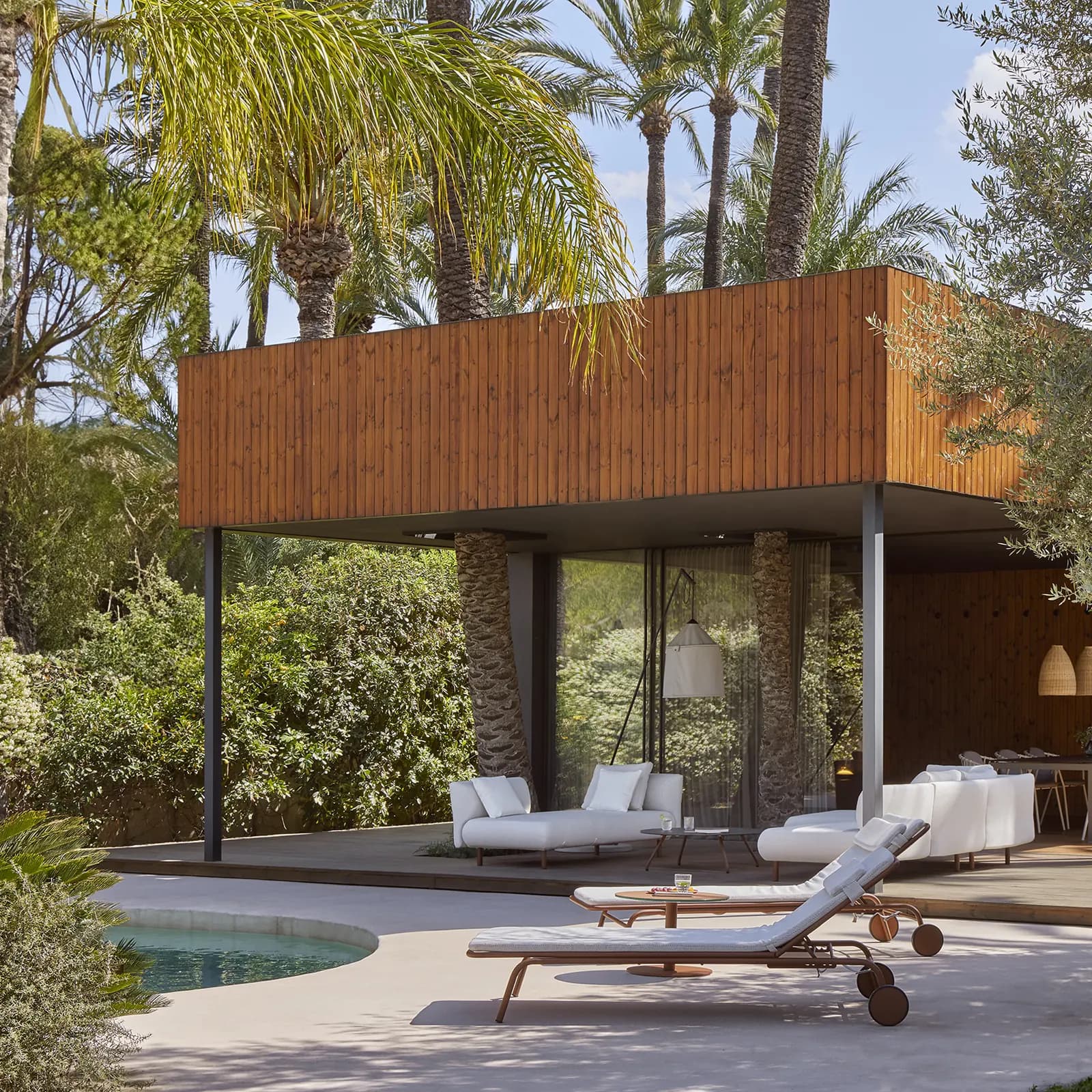 Loungers by a pool in front of a timber pavilion among palms and olive trees
